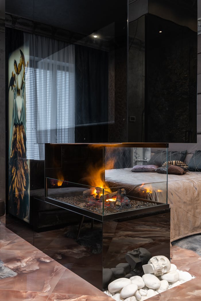 Transparent glass fireplace with bright flame placed on tiled floor with stones near bed in modern cozy bedroom at home